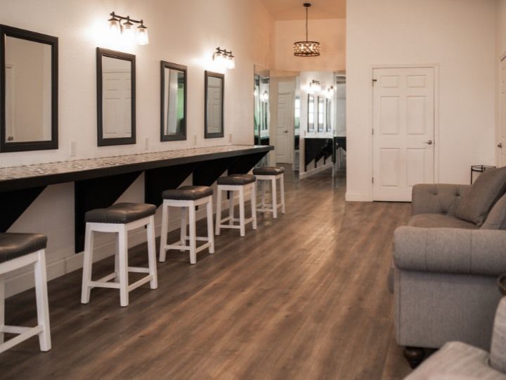 Modern salon with styling stations, mirrors, white stools, and a gray sofa on wood flooring