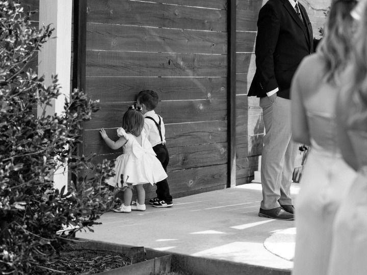 Two children standing by a large wooden door near a hedge, with adults nearby outdoors