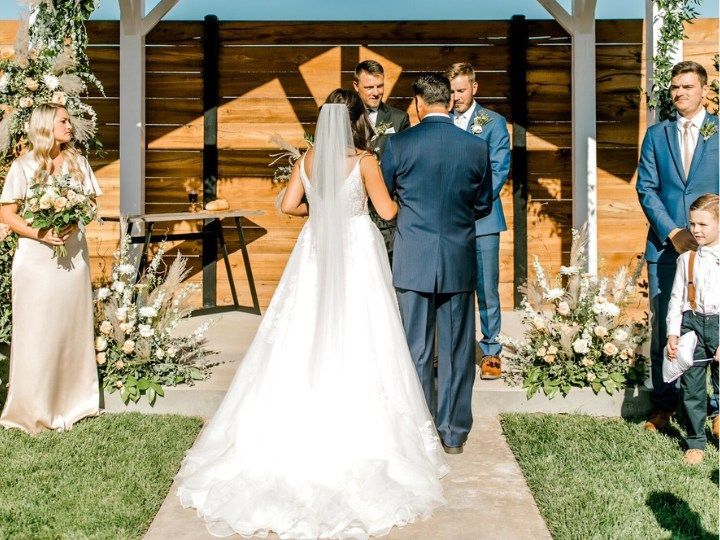Wedding ceremony under a wooden pavilion, bride in white gown facing officiant with guests nearby.