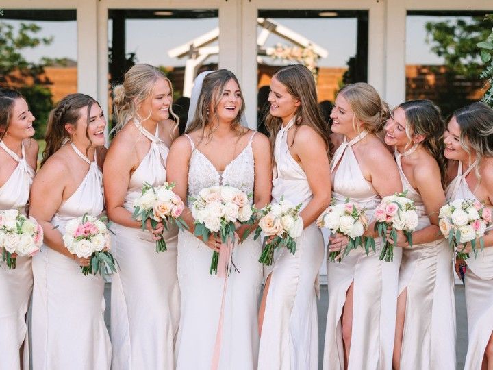 Bridesmaids in white dresses holding bouquets, smiling outdoors in front of glass doors.