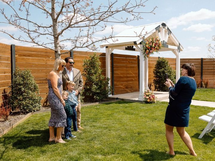 Woman taking a photo of a family standing in a grassy backyard with wooden fences and a white pergola
