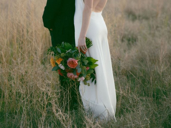 Couple standing in tall grass, bride holding a bouquet of orange and pink flowers, wedding attire