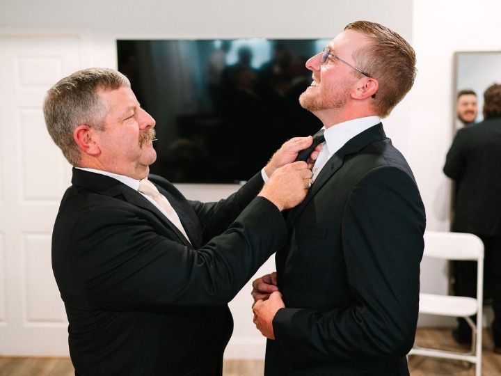 Two men in suits adjusting each other’s ties in a bright room with a TV in the background