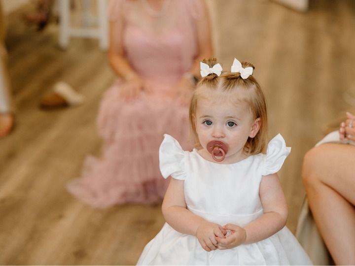 Toddler in a white dress with bows and a pacifier, sitting on a wooden floor at a party