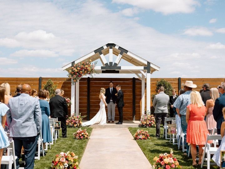 Wedding ceremony under a wooden arch by the beach, with guests seated along an aisle of flowers.