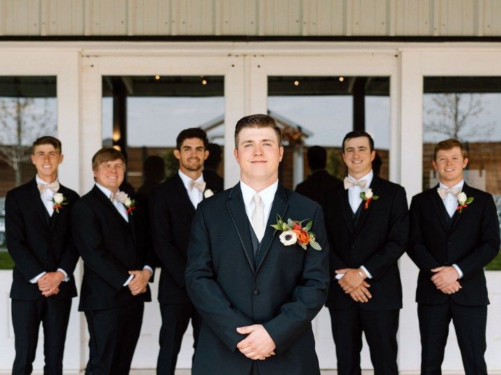 Groom and five groomsmen in black suits stand outside a building, wearing white boutonnieres.