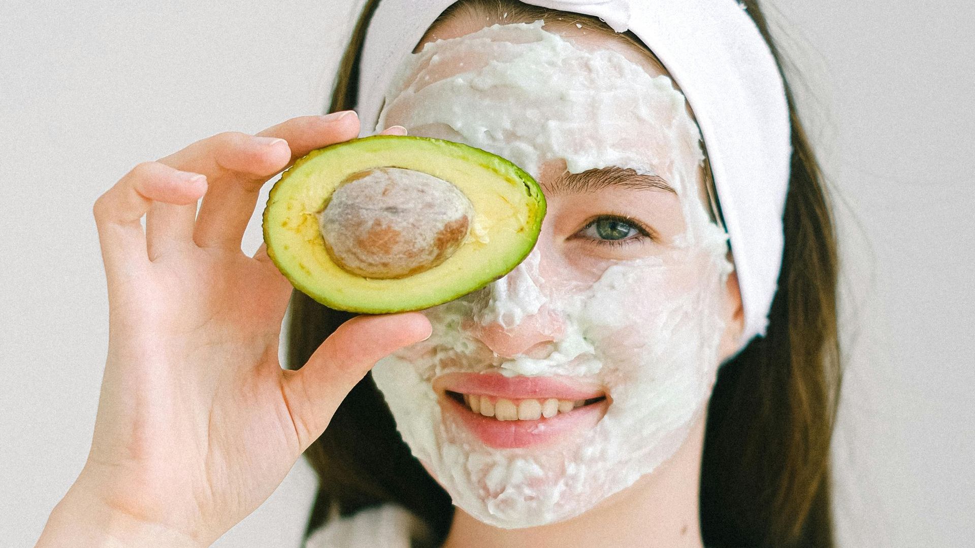 A woman with an avocado mask on her face is holding a slice of avocado in front of her eye.