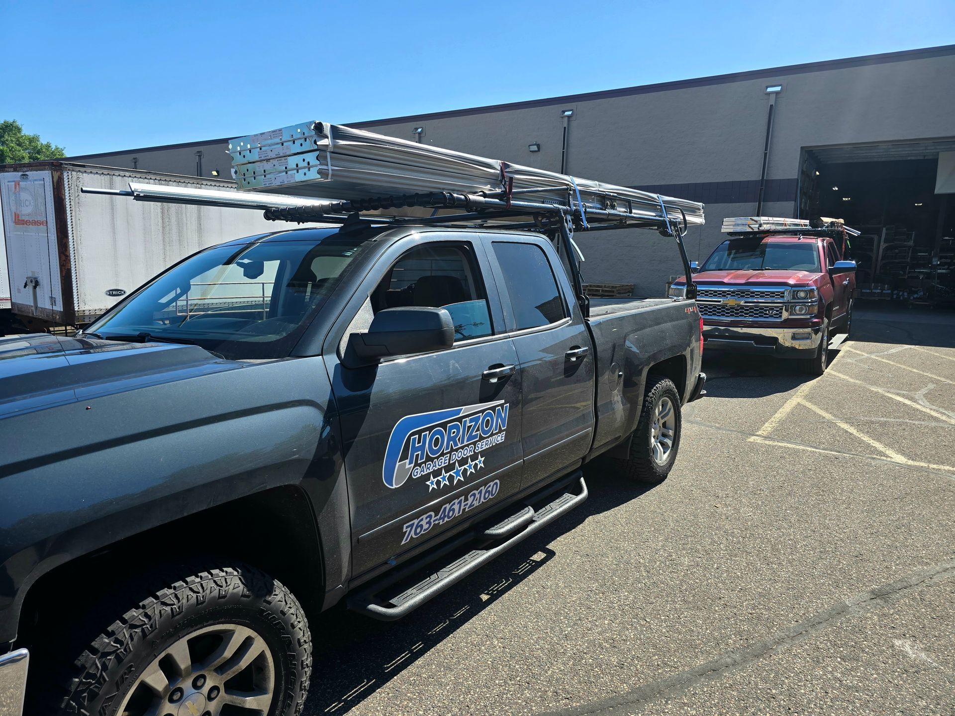 Black pickup truck with ladder rack parked in a lot, with utility vehicles behind it.