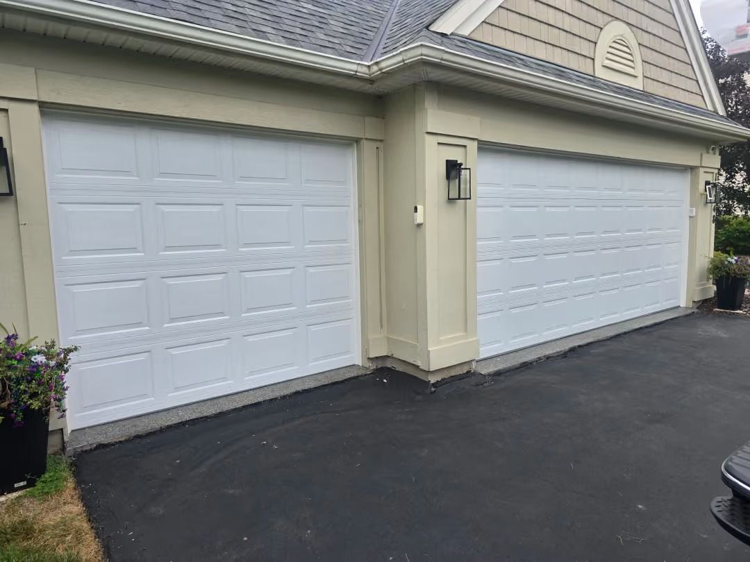 Two white garage doors on a beige house with dark driveway and small lanterns