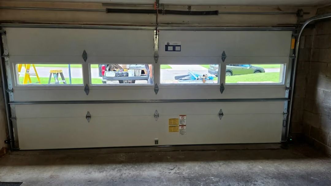 Closed white garage door with four windows and a narrow view of the outside.