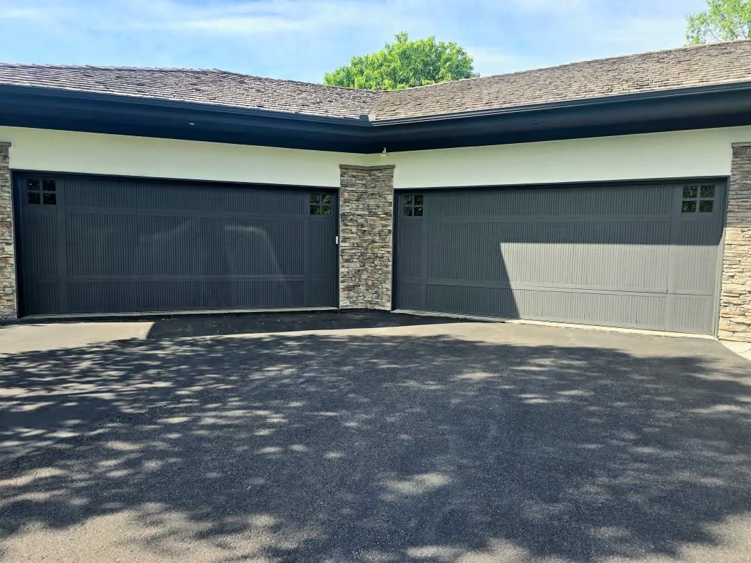 Two-car garage with dark and light doors, stone column, and sunlit driveway under a shingled roof.