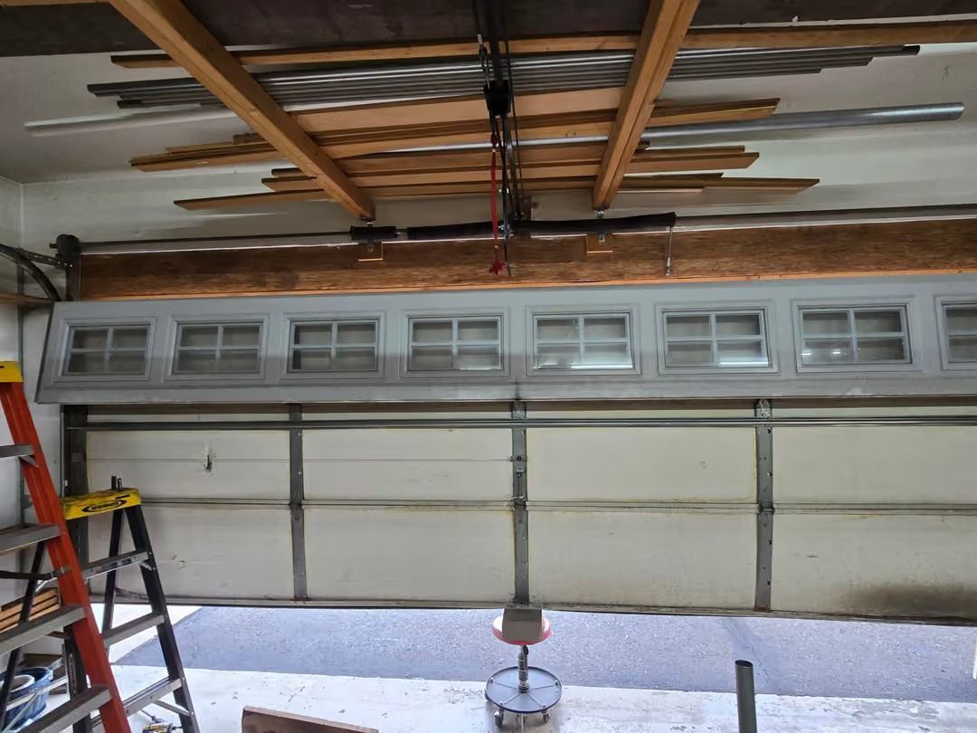 Partially open garage door with a ladder, exposed ceiling framing, and a red extension spring mechanism