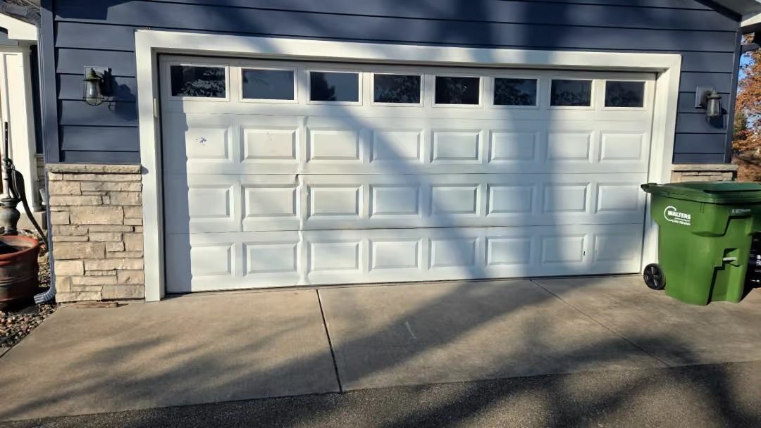 White double garage door on a blue house with stone trim, concrete driveway, and a green trash bin.