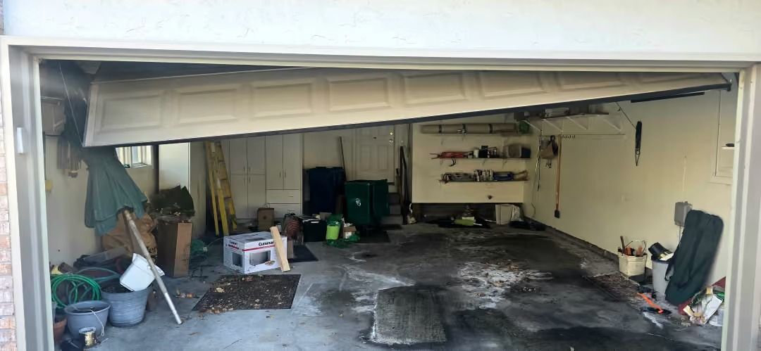 Cluttered garage interior with tools, storage shelves, and a partially open door opening onto a concrete driveway