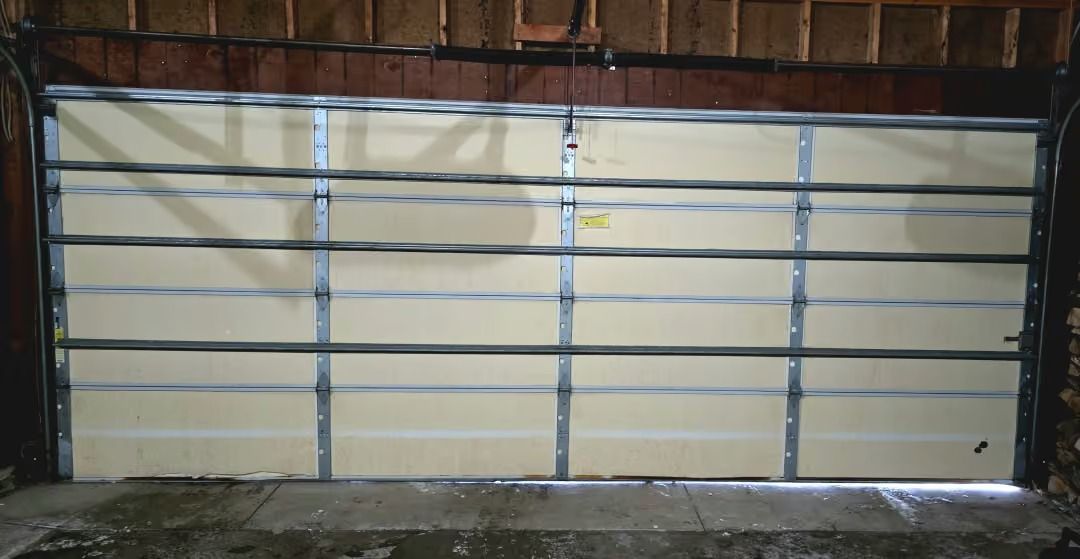 Large beige garage door with blue crossbars, viewed inside a dim workshop or garage