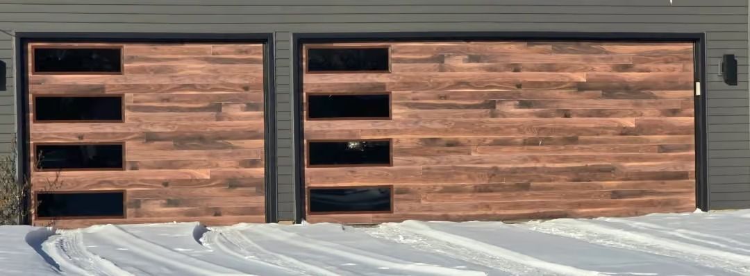 Two wooden garage doors with black windows set in a gray exterior wall, with snow piled along the bottom