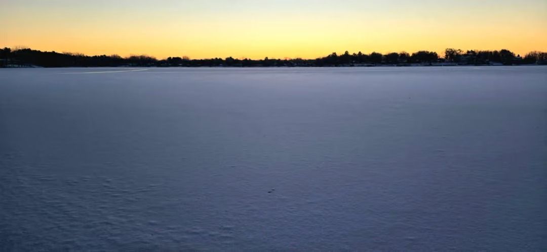 Calm frozen lake at dusk with a dark treeline silhouetted against a yellow-orange sunset sky