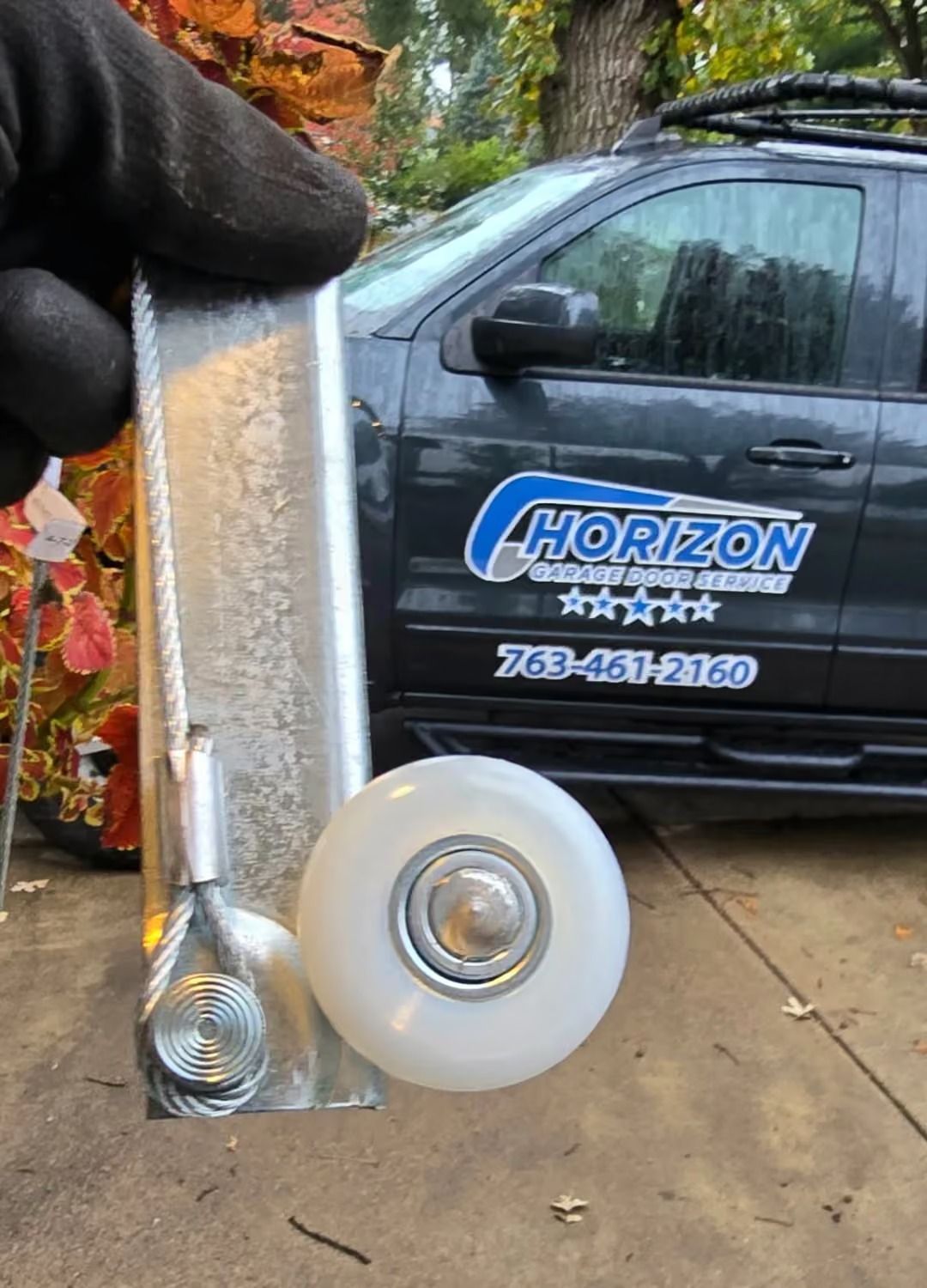 Hand holding a metal shower door roller assembly in front of a van in a wet driveway.
