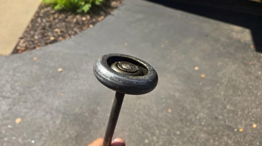 Hand holding a small black wheel or pulley on a stick over a driveway.