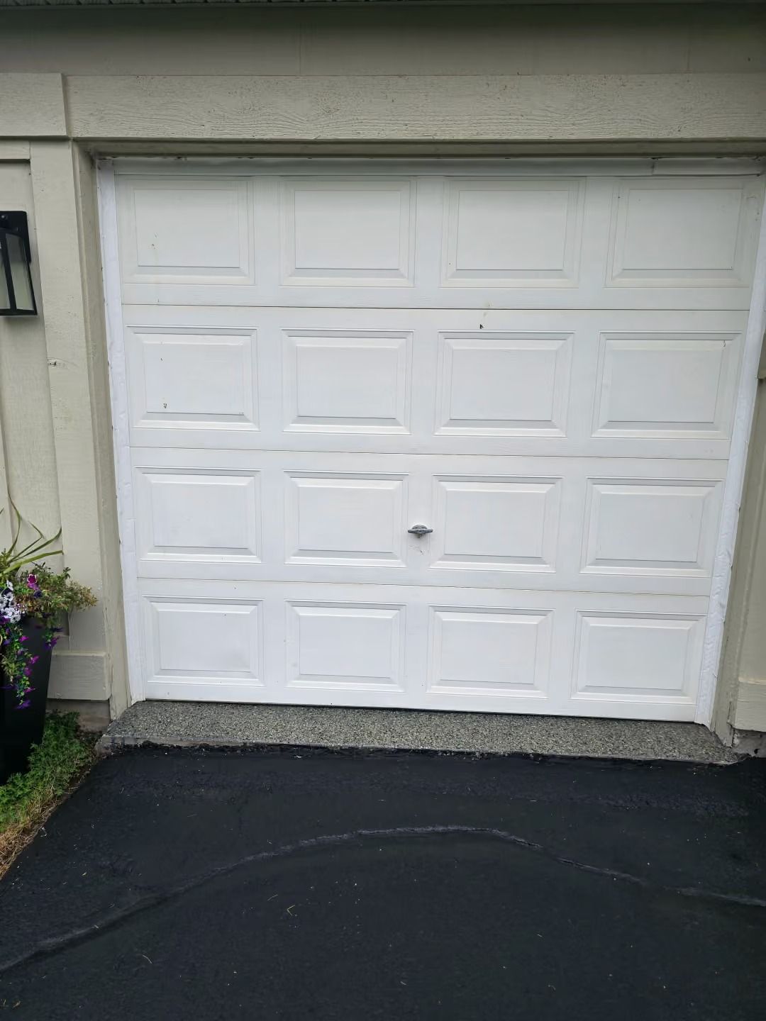 White paneled garage door on a beige house with a black driveway and flowers at the left edge
