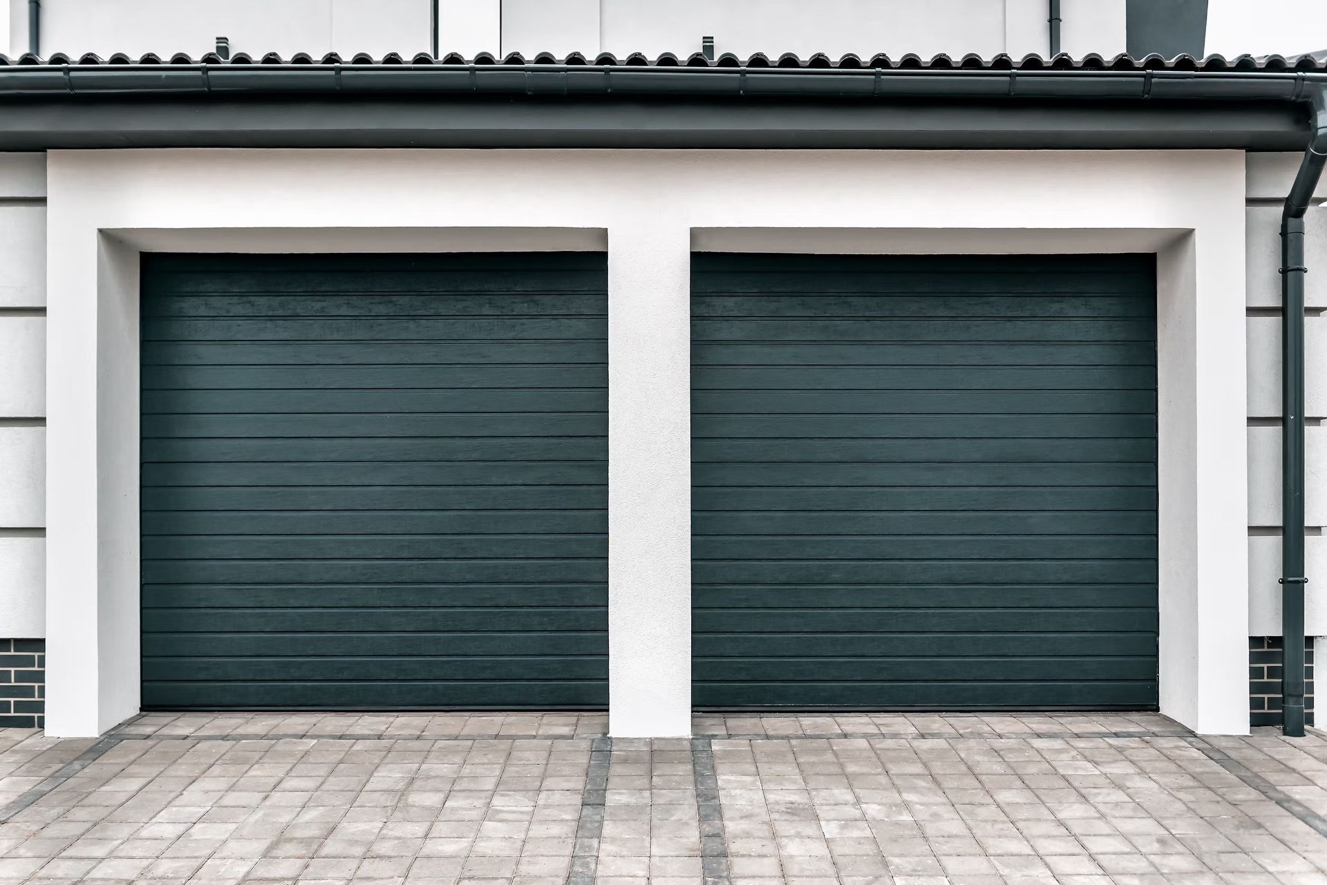 Two dark green garage doors beneath a white building facade with gray pavers in front