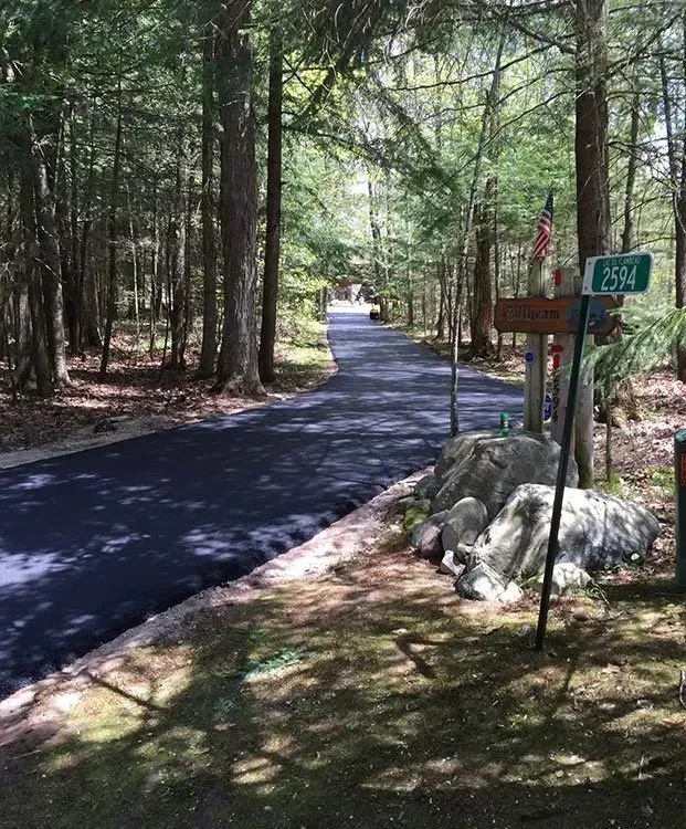 A freshly paved dark asphalt driveway curves through a sunlit wooded area, with a house number sign by a cluster of rocks. A paved driveway leads into a wooded area, past a signpost with a green house number sign and large rocks in the foreground.