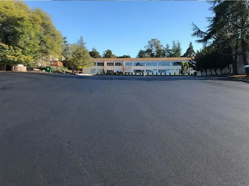 A view across a large, freshly paved black asphalt parking lot toward a modern, light-colored building surrounded by trees. A wide, freshly paved dark asphalt parking lot leading up to a beige, multi-story commercial building under a clear sky.