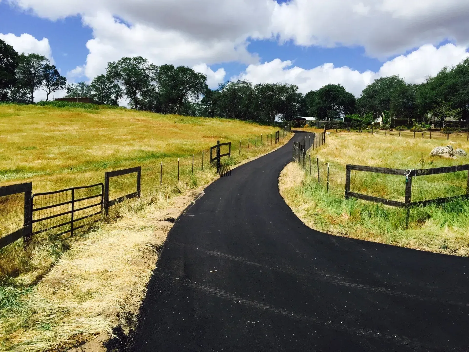 A freshly paved black asphalt driveway winds through a rural landscape with golden grassy hills and wooden fences.A freshly paved black asphalt driveway winds through a rural landscape with golden grassy hills and wooden fences. A paved driveway curves through a rural landscape with dry, golden fields bordered by wooden fences under a cloudy sky.