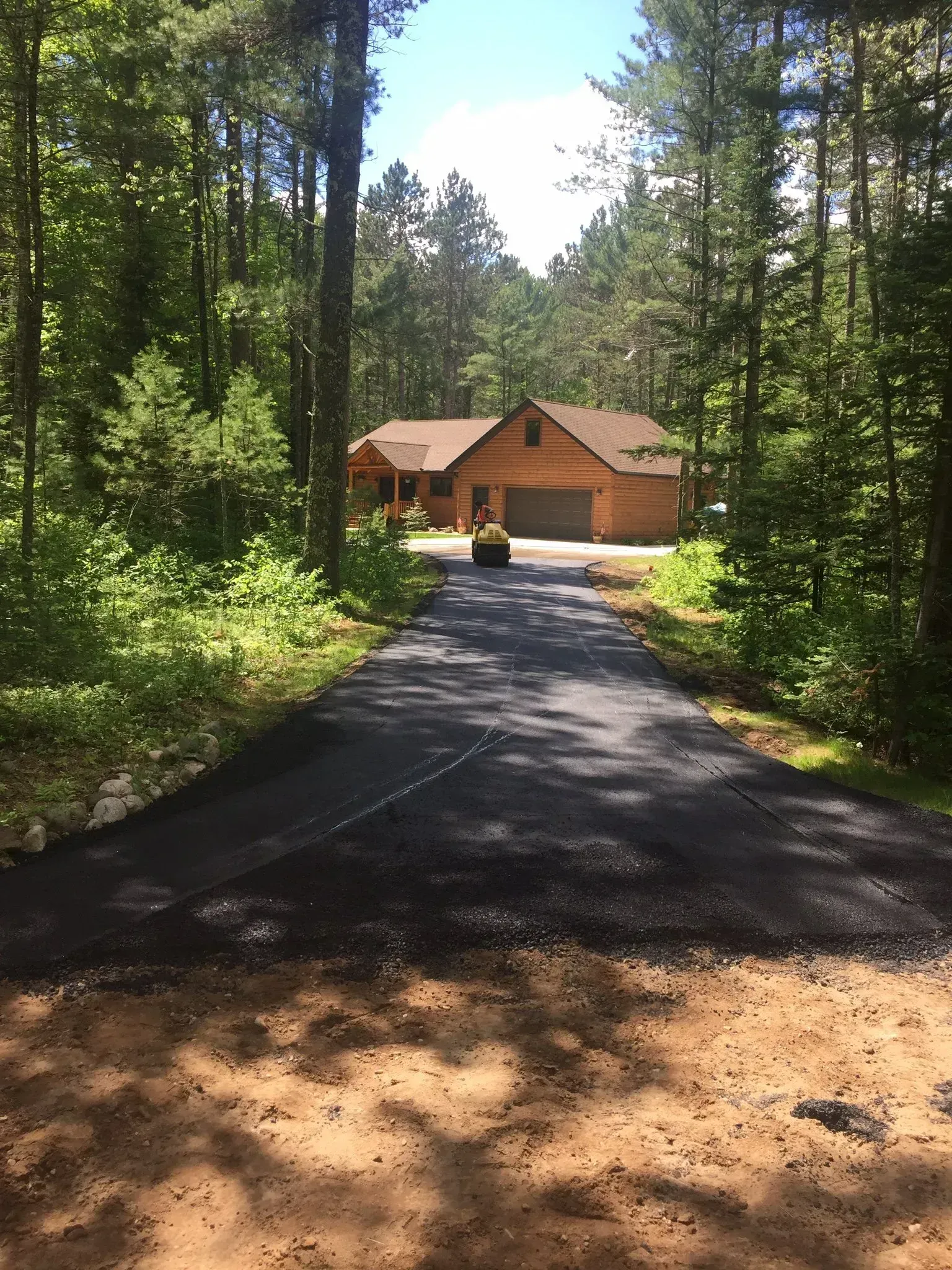 A freshly paved asphalt driveway leading through a wooded area to a single-story house with a wooden exterior. A newly paved asphalt driveway leads through a wooded area toward a brown, one-story house.
