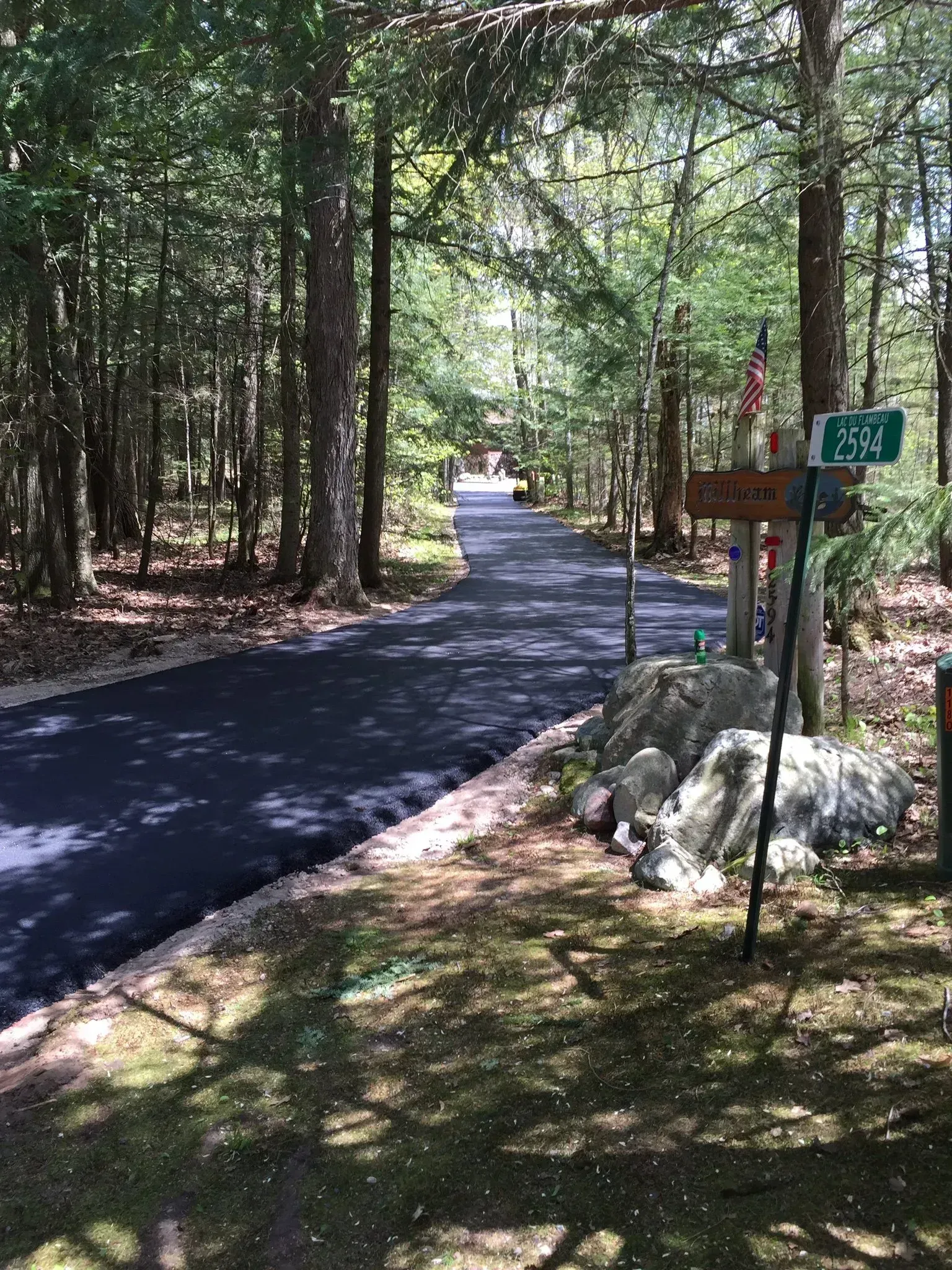 A freshly paved asphalt driveway winds through a sunny, wooded area, past large rocks and a small directional sign. A freshly paved driveway leads into a sun-dappled forest, past large boulders and a green address sign.