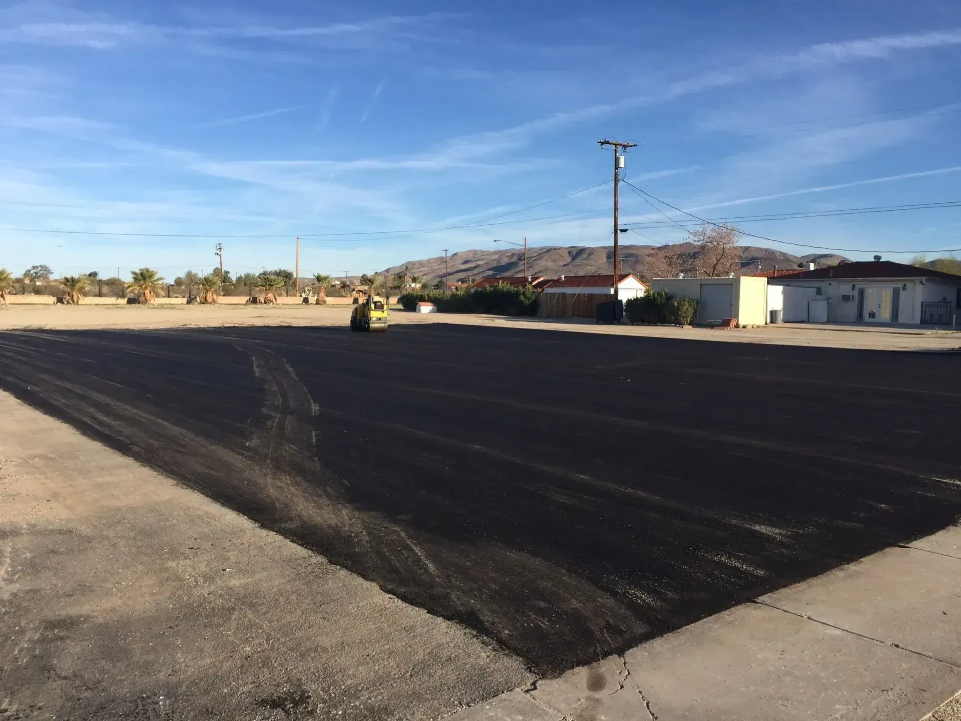 A paving machine works on a patch of fresh, dark asphalt in a bright, sunny outdoor lot with distant mountains. A yellow steamroller compacts fresh, dark asphalt on a large lot under a clear blue sky, with buildings in the distance.
