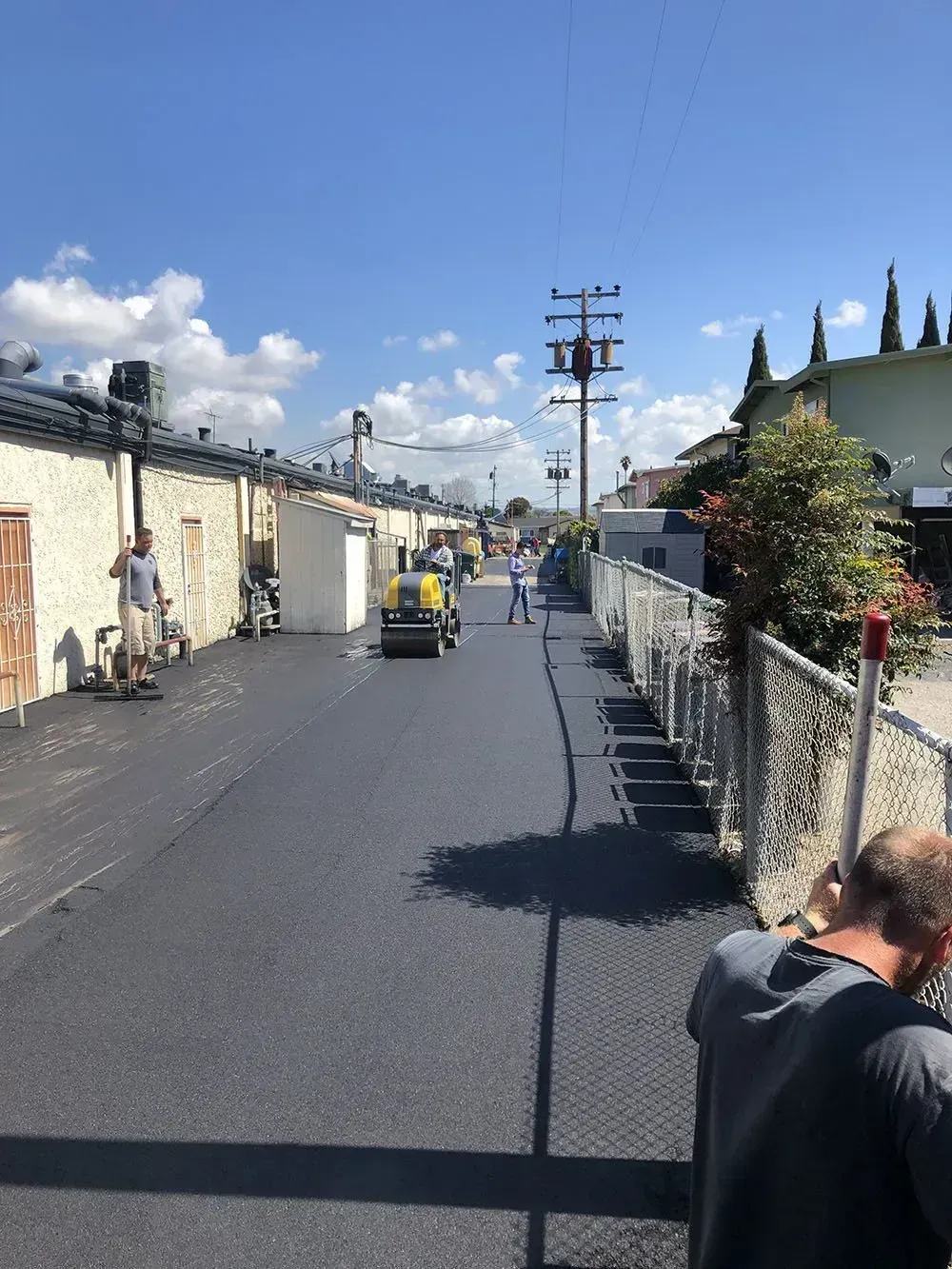 A construction worker stands in the foreground, looking toward a roller compacting new asphalt on a narrow alleyway. Workers pave a narrow alleyway with fresh asphalt using a steamroller on a sunny day.