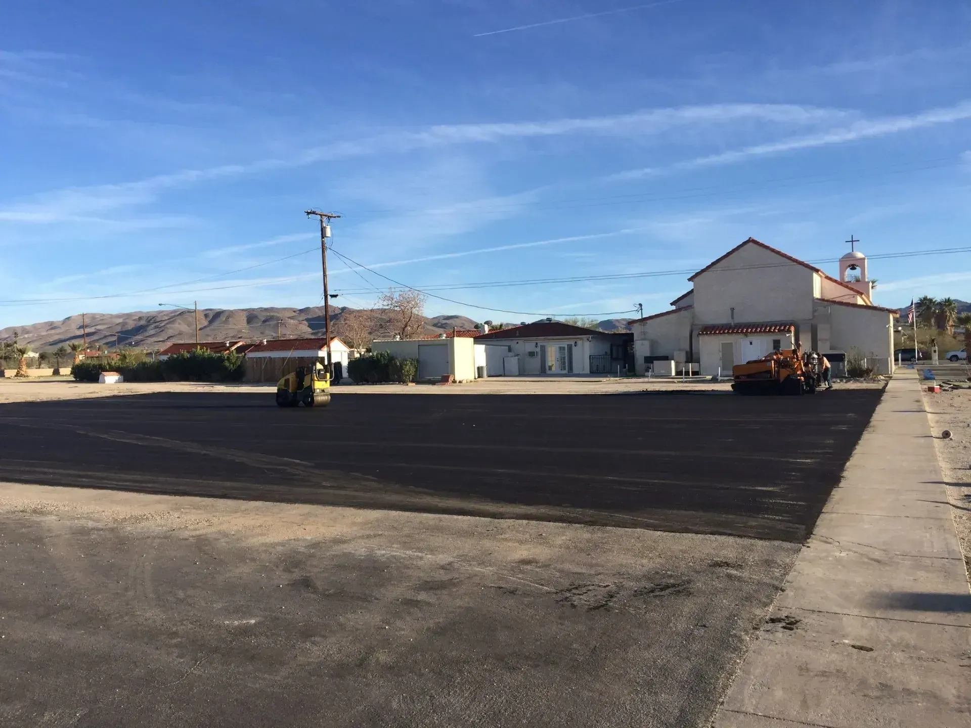 A view of a parking lot being resurfaced with fresh black asphalt, with construction rollers and a nearby church building. Construction workers use heavy rollers to pave a large asphalt parking lot in front of a white church building.