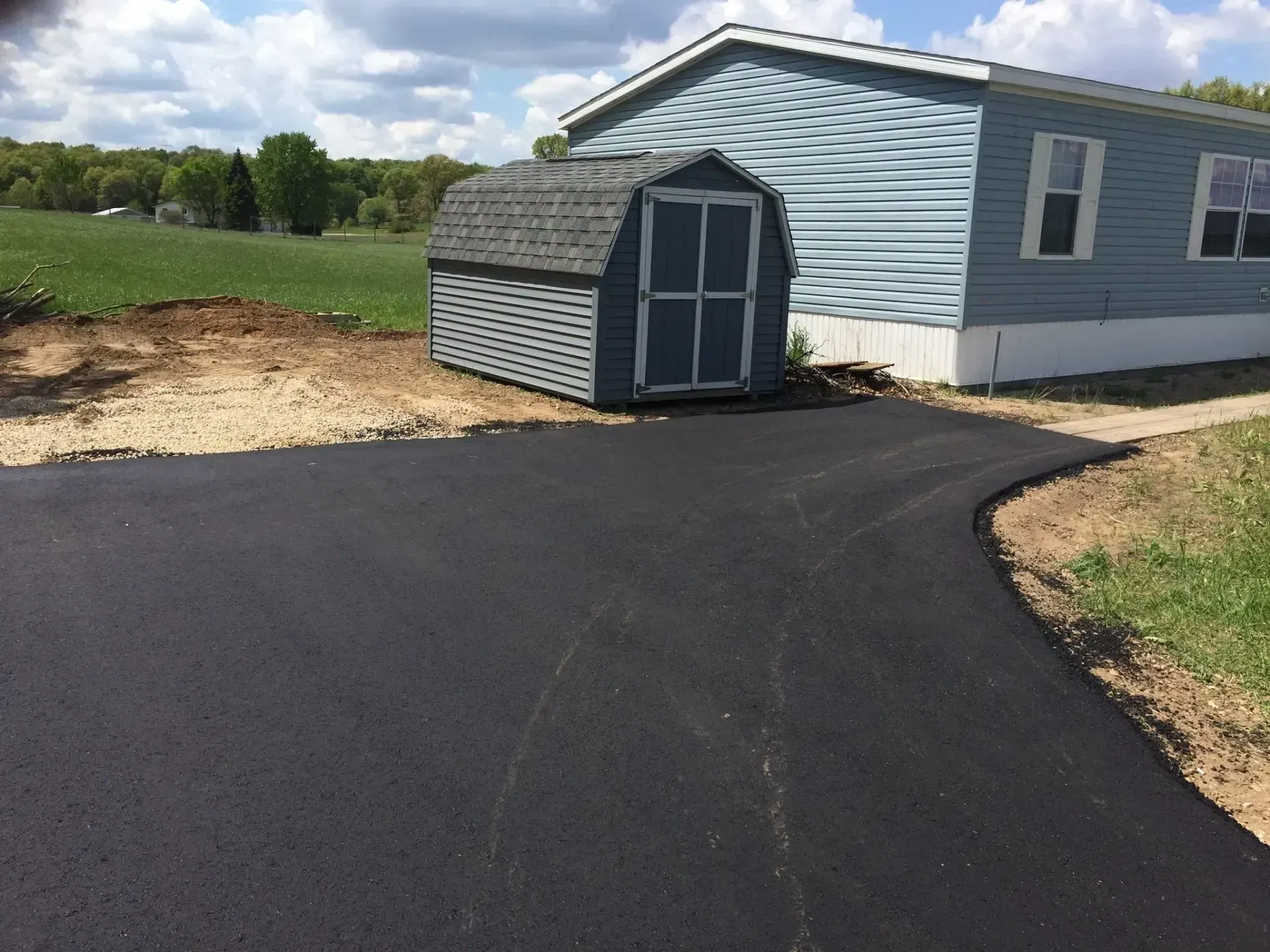 A new black asphalt driveway curves past a small blue storage shed and the side of a blue manufactured home. A newly paved asphalt driveway leads toward a blue storage shed next to a light blue mobile home in a grassy yard.