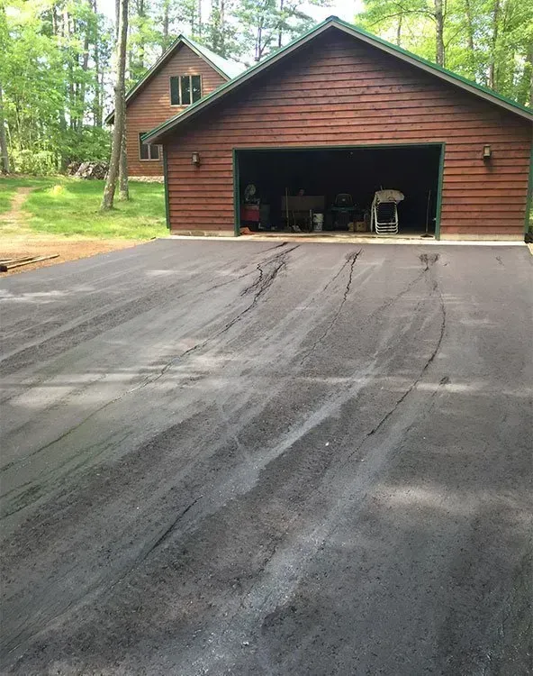 A freshly paved, dark asphalt driveway showing tire track indentations leading toward a wooden garage and house. A wooden garage with an open door stands behind a newly paved asphalt driveway featuring prominent tire tracks.