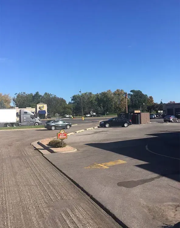 An asphalt parking lot under a clear blue sky, featuring parked cars, a semi-truck in the distance, and a small sign. A parking lot on a sunny day with cars and a semi-truck visible in the background.