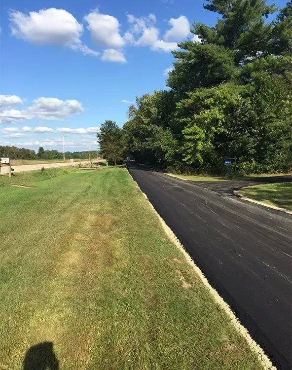 A freshly paved black asphalt road curves past a green grass verge and a dense line of trees under a blue, cloudy sky. A freshly paved black asphalt road runs alongside a bright green grassy field under a sunny blue sky with scattered clouds.