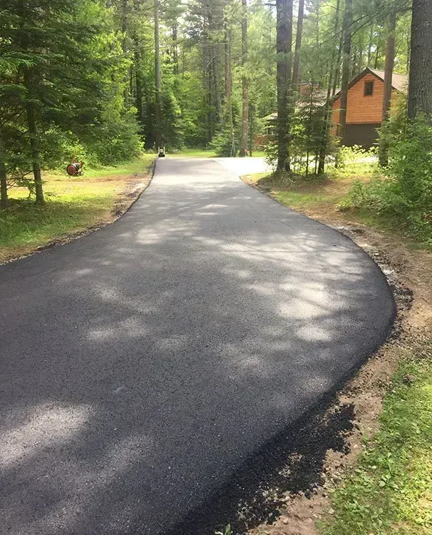 A newly paved asphalt driveway curves through a wooded area toward a house in the distance. A freshly paved black asphalt driveway curves through a sunlit wooded area toward a house in the distance.