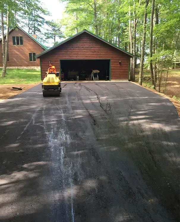 A yellow steamroller compacts fresh black asphalt on a driveway in front of a wooden garage and house in a wooded area. A yellow steamroller compacts fresh black asphalt on a driveway in front of a wooden garage and a house in the woods.