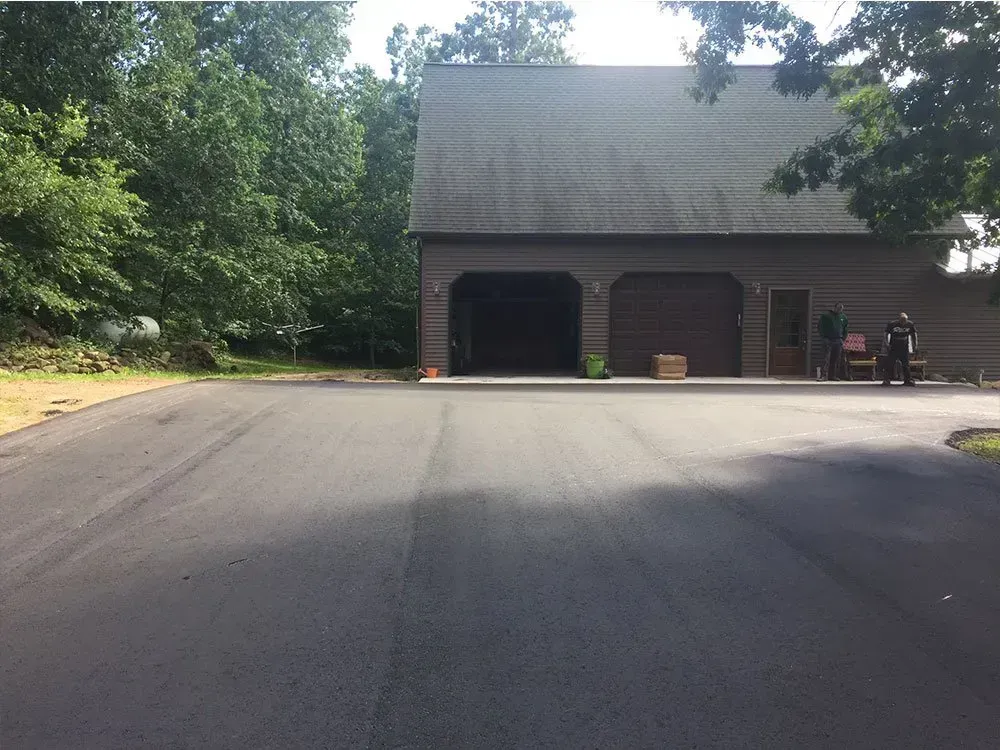 A freshly paved asphalt driveway leading to a two-story garage with a dark roof surrounded by green trees. A newly paved asphalt driveway leads to a large, two-bay garage nestled in a wooded area.