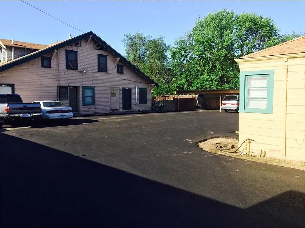 A paved parking lot between a light-pink two-story building and a tan structure under a clear blue sky. A paved lot between a two-story pink residential building and a smaller tan structure with a light blue window frame.