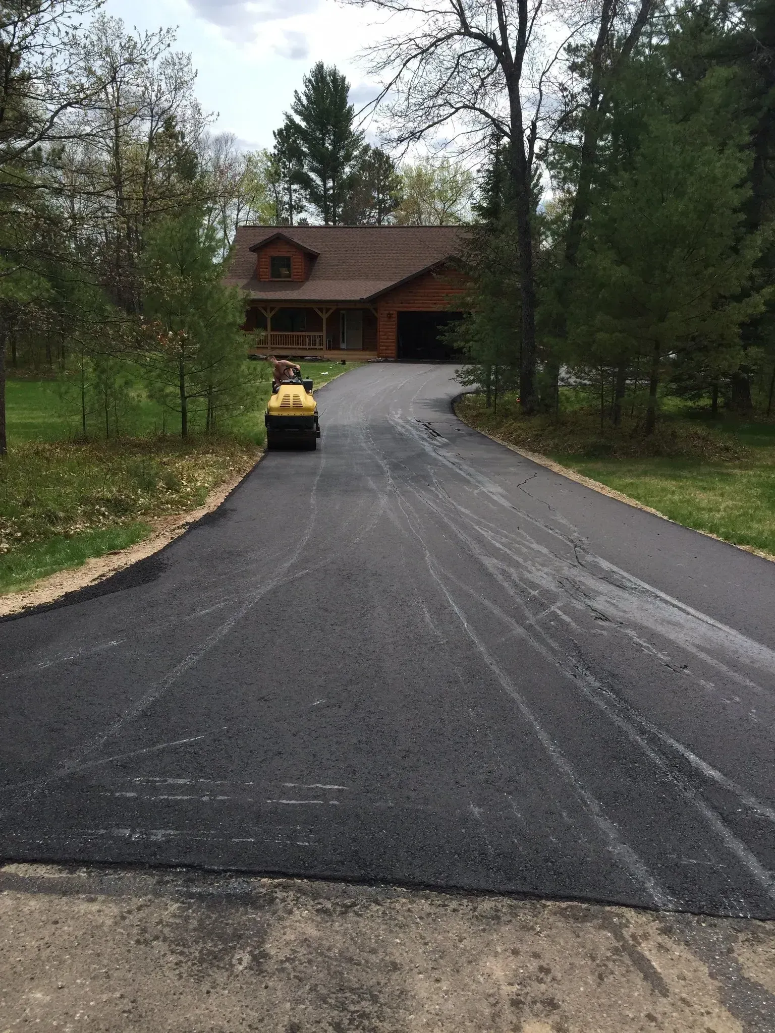 A yellow steamroller compacting a freshly paved asphalt driveway leading to a house surrounded by trees. A yellow steamroller compacts fresh, dark asphalt on a driveway leading to a house surrounded by trees.