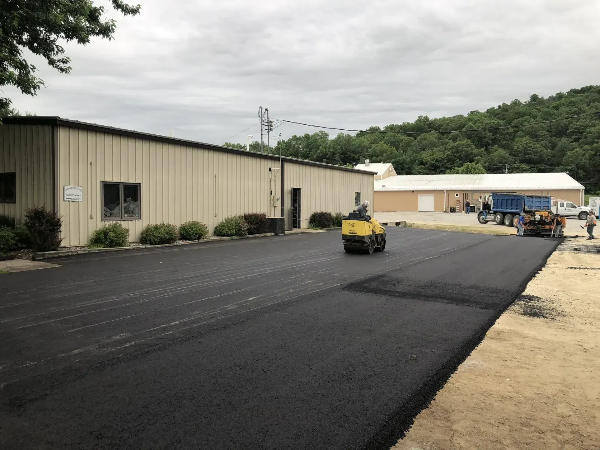 A roller compacts fresh black asphalt on a driveway alongside industrial buildings under an overcast sky. A yellow asphalt roller compacts a freshly paved black parking lot next to a tan commercial building.