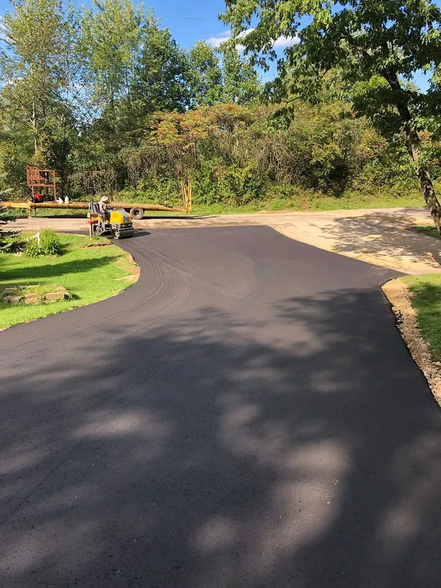 A newly paved black asphalt driveway curves through a lush green yard towards a wooded area. A freshly paved black asphalt driveway winding through a green, tree-lined residential property on a sunny day.