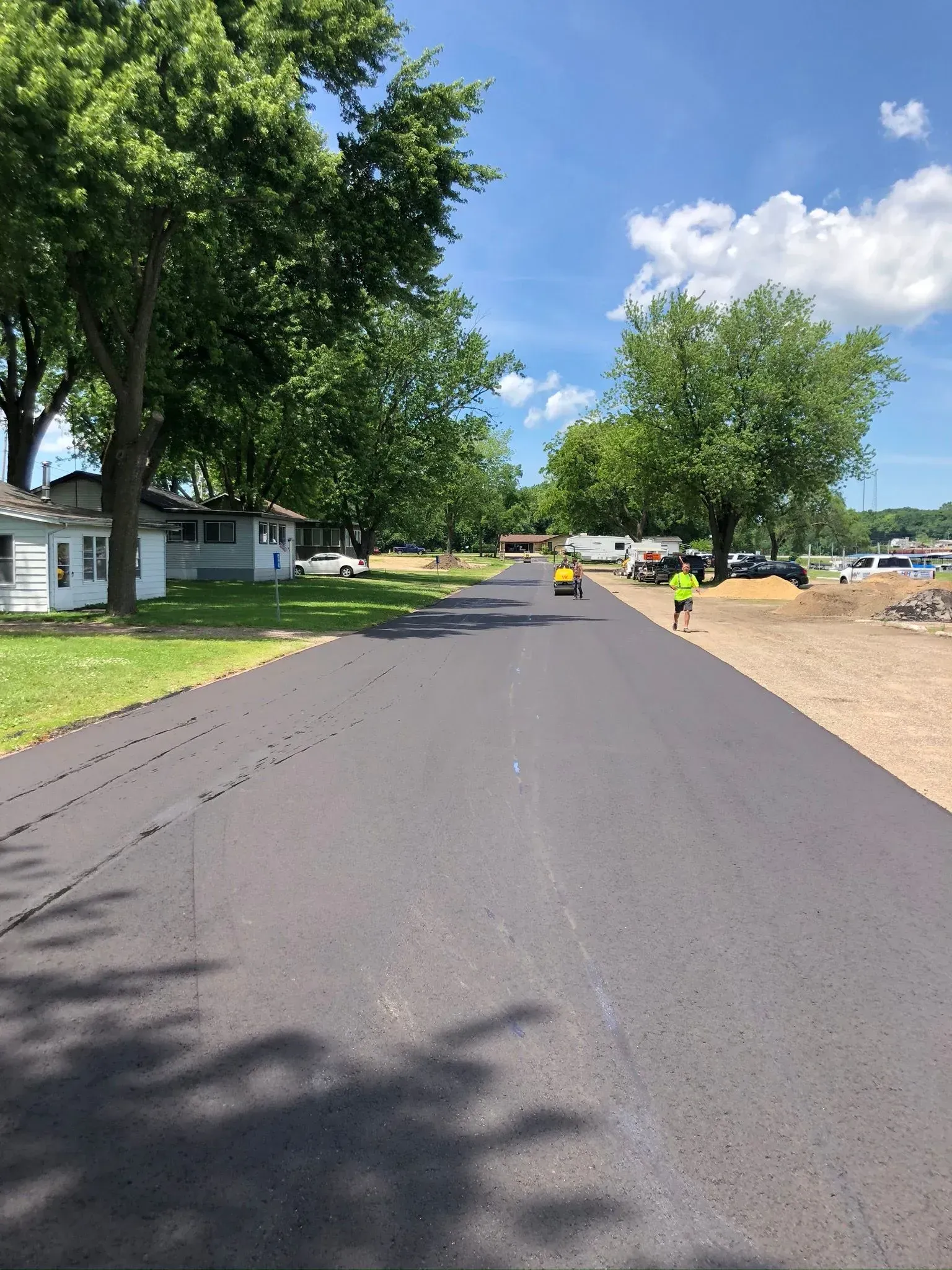 A newly paved black asphalt road stretches past houses and trees with a construction roller and worker in the distance. A freshly paved asphalt road under a bright blue sky, with trees on the left, a worker on the right, and a roller ahead.