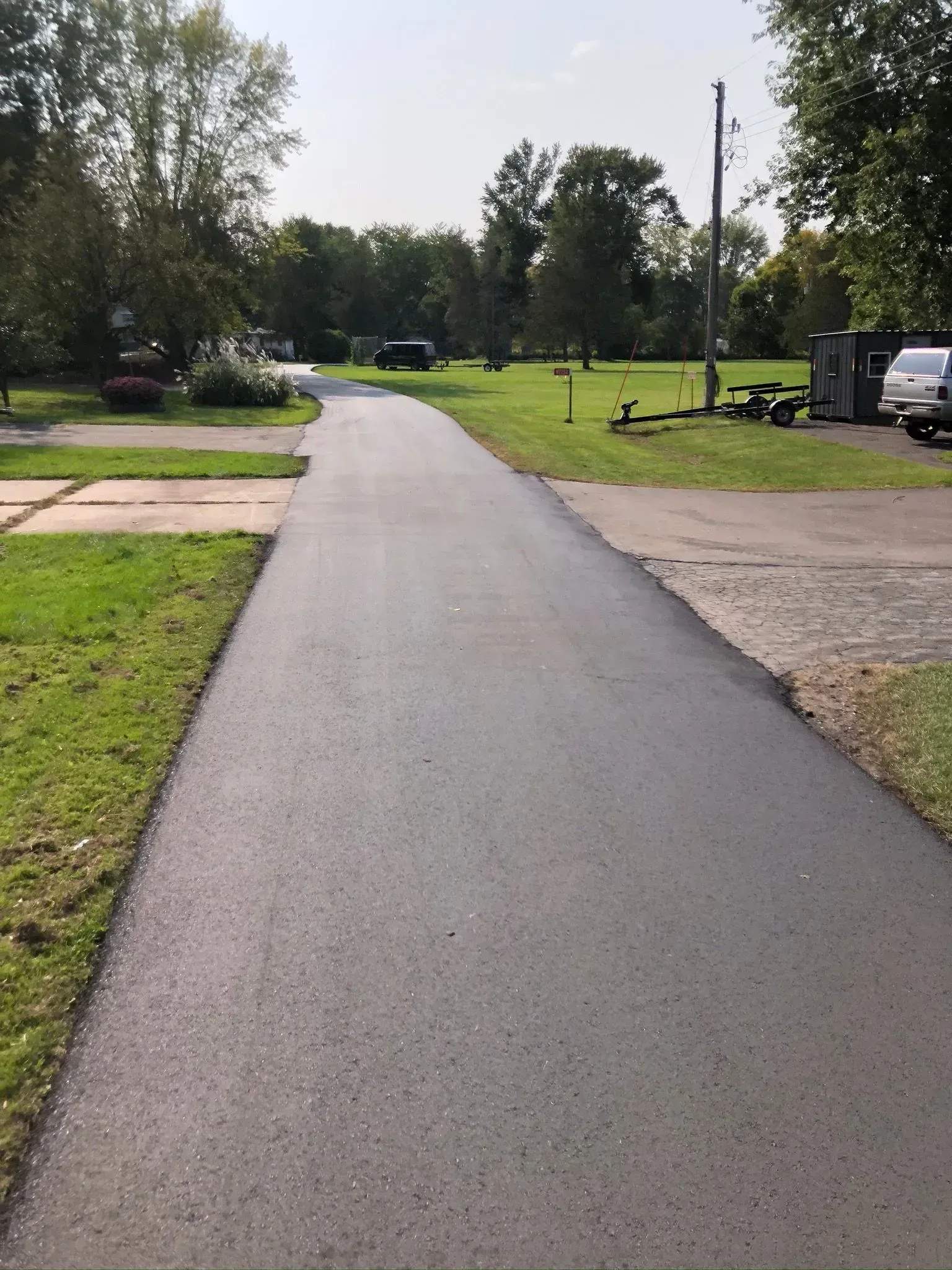 A freshly paved, dark gray asphalt path extends through a sunny outdoor setting with grass and trees on either side. A freshly paved black asphalt driveway stretches through a grassy area toward trees under a clear sky.