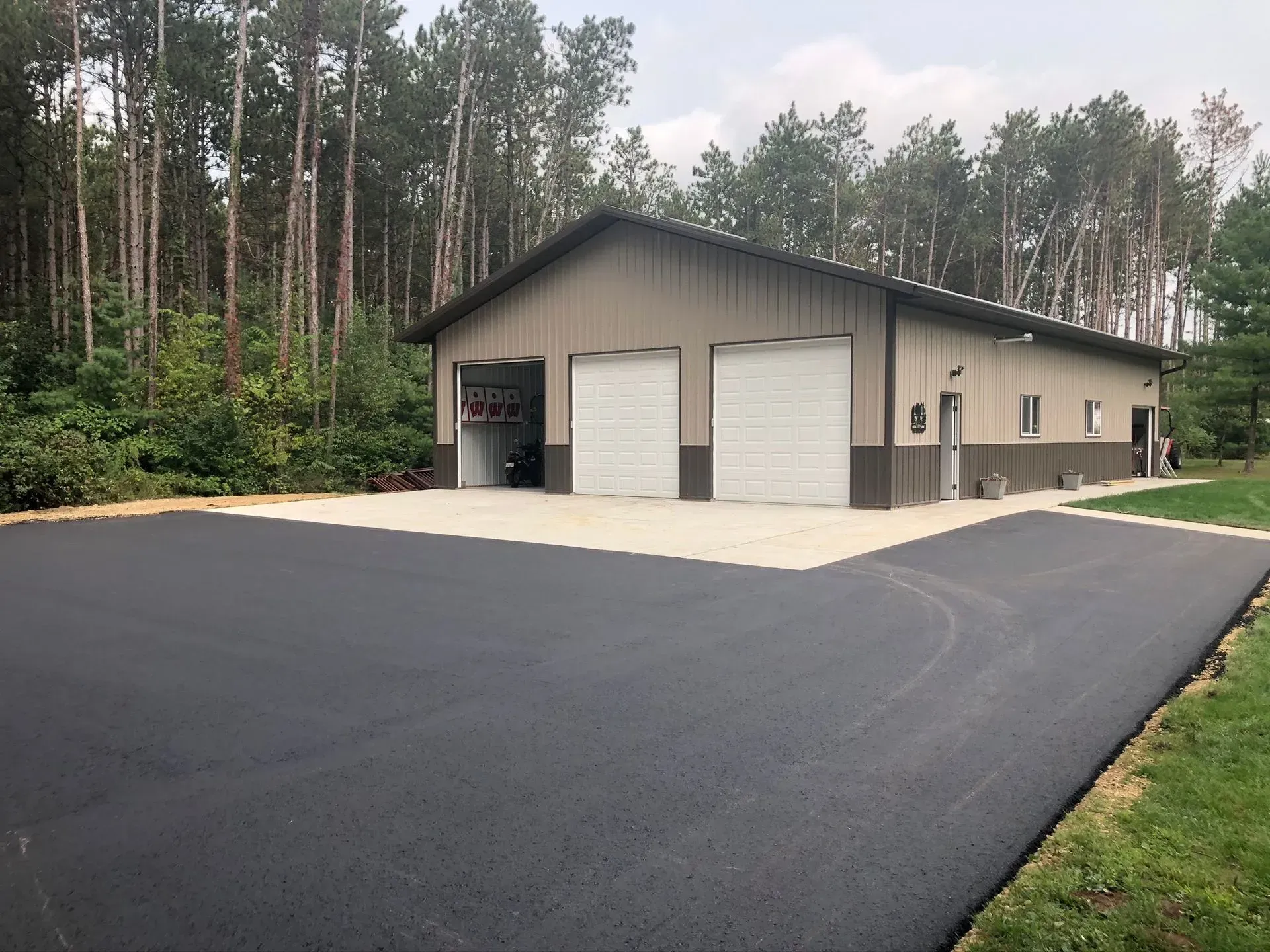 A tan metal outbuilding with two white garage doors and a large, freshly paved black asphalt driveway. A tan, metal-sided building with two white garage doors, set on a large, freshly paved asphalt driveway near a forest.