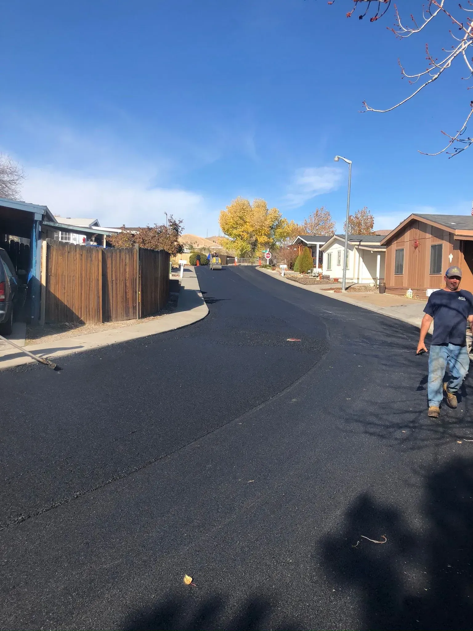A person walks beside a freshly paved asphalt road winding through a residential street on a sunny day. A worker stands on a freshly paved black asphalt road in a residential area on a sunny day.