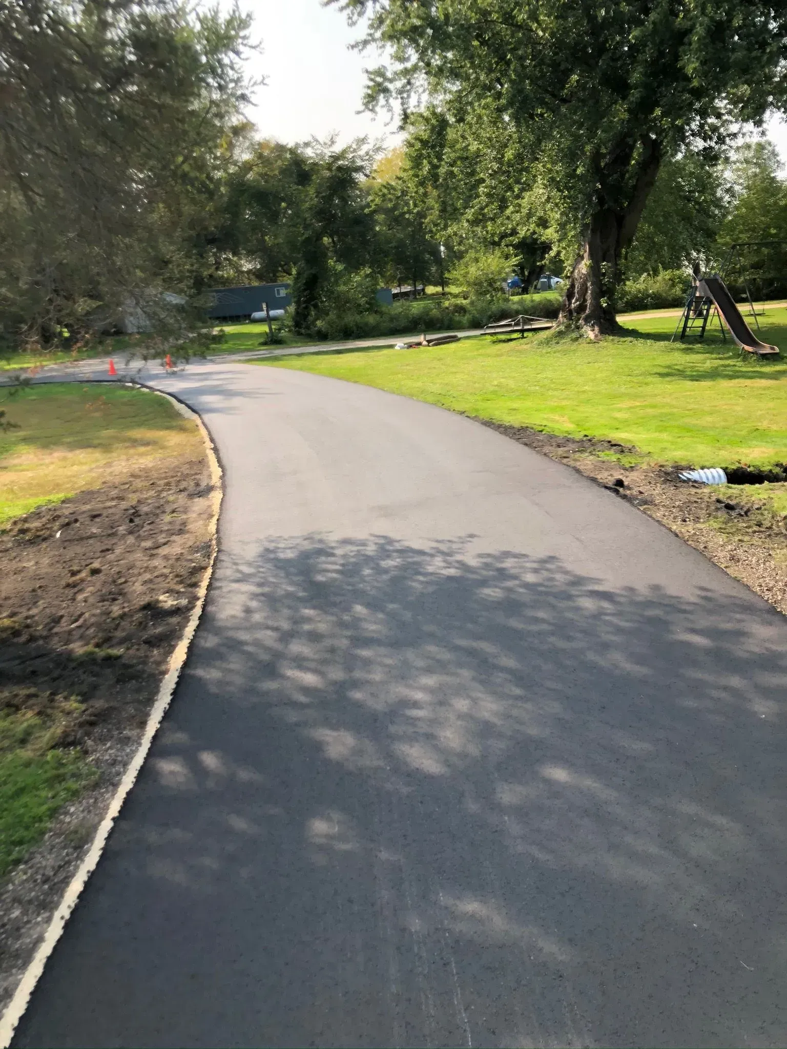 A newly paved asphalt driveway curves through a residential lawn toward a large tree. A newly paved asphalt driveway curves through a grassy yard toward trees on a sunny day.