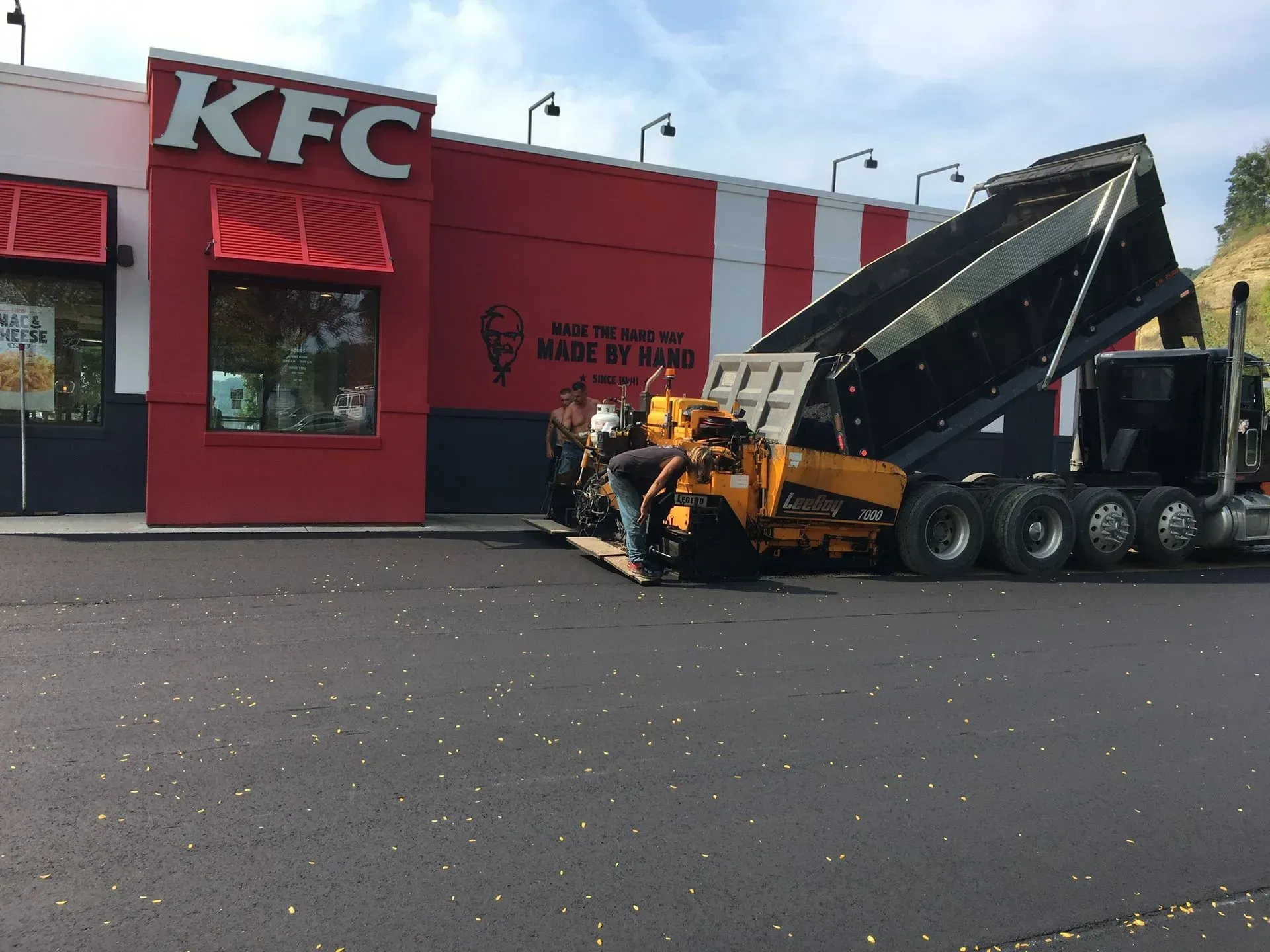 A yellow asphalt paving machine is depositing hot mix in front of a KFC restaurant, with a dump truck tilted to load it. A yellow construction paving machine works on a freshly laid black asphalt parking lot in front of a KFC restaurant.